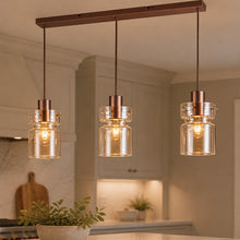 Load image into Gallery viewer, Three-light copper-finished pendant ceiling fixture with clear glass shades hanging over a kitchen island, with warm glowing bulbs and a softly blurred kitchen background.

